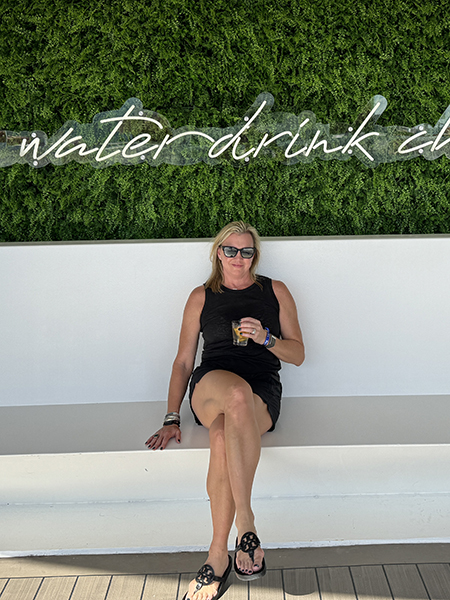 Amy Westerman with a drink in front of a wall that says "Save water drink champagne"