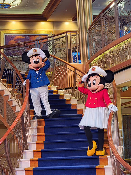photo on board a Disney Cruise Line ship, of Mickey and Minnie Mouse wearing captain's hats and saluting, standing on a staircase aboard one of their ships.