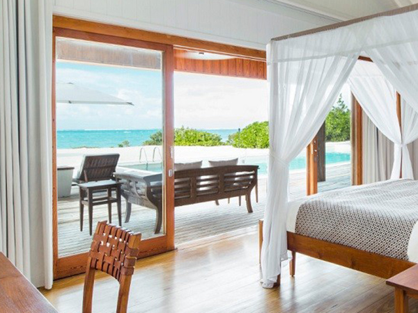 photo of a bedroom in a luxury villa from Villas of Distinction, showing a view of the ocean in a tropical setting, with a bed and desk, as well as a wall of windows/doors 