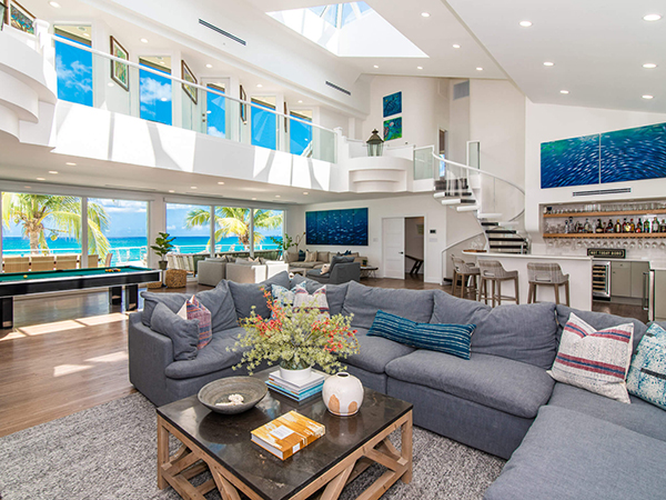 photo of living area of a villa in a tropical location, from Villas of Distinction. High ceilings, large sofa with coffee table, pool table, bar, numerous windows looking over a beach, with a spiral staircase and skylight 