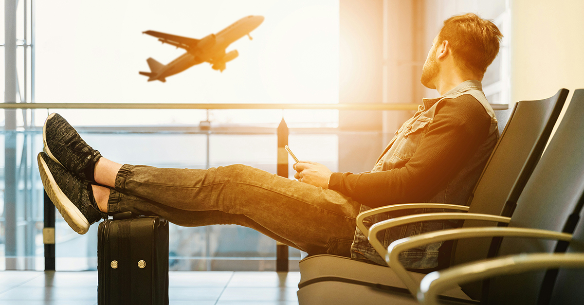 photo of a young man in the airport, with his legs resting on his carryon bag, looking out the window as an airplane takes off outside